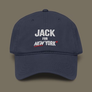 May include: Navy blue baseball cap with the white text "JACK" above "FOR NEW YORK". The word "NEW YORK" is in white with a red underline. The hat is made of a soft material and has a curved brim.