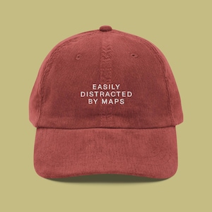 May include: A burgundy corduroy baseball cap with white embroidered text that reads "EASILY DISTRACTED BY MAPS".