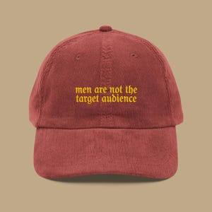以下が含まれることがあります： 黄色い文字で「men are not the target audience」と刺繍された、マルーン色のコーデュロイベースボールキャップ。 キャップはカーブしたつばと丸みを帯びたトップを備えています。