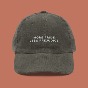 May include: A dark green corduroy baseball cap with the text "More Pride Less Prejudice" embroidered in white on the front.