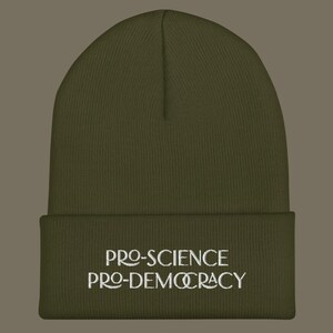 May include: An olive green cuffed beanie featuring white embroidered text on the front that reads PRO-SCIENCE PRO-DEMOCRACY. The knit hat has a ribbed texture and a classic rounded crown shape, providing a simple and functional head covering.