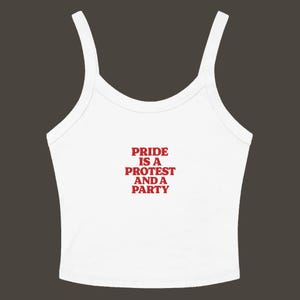 Puede incluir: Camiseta sin mangas blanca con tirantes finos. El texto rojo en la parte delantera dice "PRIDE IS A PROTEST AND A PARTY". La parte superior está hecha de un material suave y es adecuada para ropa casual.
