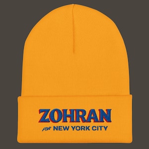 Könnte beinhalten: Senfgelbe Mütze mit dem Namen "ZOHRAN" in Blau mit rotem Schatten gestickt und dem Text "FOR NEW YORK CITY" darunter. Die Mütze ist aus einem weichen, gestrickten Material.