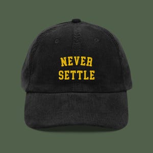 May include: Black corduroy baseball cap featuring the text "NEVER SETTLE" in yellow block lettering. The cap has a curved peak and a rounded crown. A fashionable accessory for casual wear.