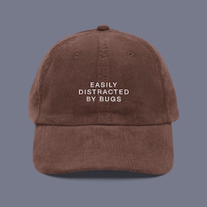 Puede incluir: Una gorra de béisbol de pana marrón con bordado blanco que dice "EASILY DISTRACTED BY BUGS".