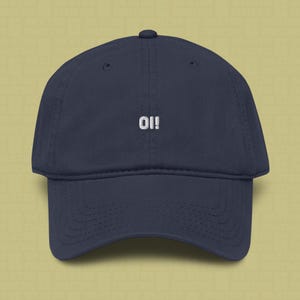 May include: A navy blue baseball cap featuring the word "Oi!" embroidered in white thread on the front centre. The cap has a structured crown, a curved brim, and a button at the top centre, showcasing a clean and simple design.