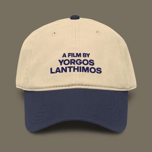 May include: A beige and navy blue baseball cap. The cap features the text "A FILM BY YORGOS LANTHIMOS" in navy blue lettering across the front. The cap has a curved brim and a button on top.