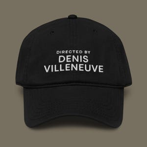May include: Black baseball cap with the text "DIRECTED BY DENIS VILLENEUVE" embroidered in white. The cap has a curved brim and a rounded crown. A stylish accessory for film enthusiasts.