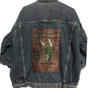 Puede incluir: Chaqueta vaquera azul descolorida. La parte trasera presenta un parche rectangular con las palabras "RED HOT CHILI PEPPERS" y un gráfico de diablo verde. La palabra "NIRVANA" también es visible en el parche.
