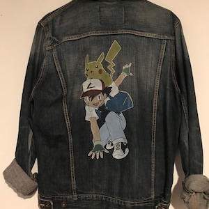 Könnte beinhalten: Eine blaue Jeansjacke mit einem handgemalten Bild eines Zeichentrickcharakters aus der Anime-Serie Pokemon. Der Charakter trägt ein blau-weißes Hemd und eine rot-weiße Baseballkappe. Der Charakter reitet auf dem Rücken eines gelben Pikachu.
