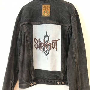 Puede incluir: Chaqueta vaquera negra con un parche blanco con el nombre de la banda "Slipknot" en letras marrones. La chaqueta tiene una etiqueta marrón con el texto "Auntie Flusso" en la parte posterior.