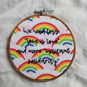 Op de afbeelding: Een ronde borduurring met een stoffen ontwerp met regenboogbogen en de tekst "We need less 'love is love' and more 'militant solidarity'". De ring is gemaakt van hout en heeft een metalen spanmechanisme.