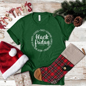 May include: Green t-shirt with a white circle graphic that says "black friday" with a list of activities around the text. The shirt is on a white wood background with a red Santa hat, a small gift box, and a red and black plaid stocking.