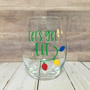Puede incluir: Una copa de vino de vidrio transparente con el texto "Let's get lit" en letras verdes y una cadena de luces navideñas de colores en el lateral.