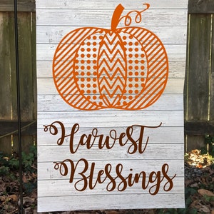 May include: A rectangular garden flag with a white, weathered wood effect background. An orange pumpkin design is centered on the flag, with the words "Harvest Blessings" in a brown script below. The pumpkin has a variety of patterns.