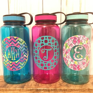 May include: Three reusable water bottles with personalized monograms. The bottles are turquoise, pink, and teal. The monograms are in a circle with a patterned background. The monograms are "AHN", "T", and "E".
