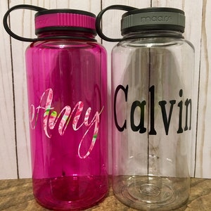 May include: Two clear plastic water bottles with black lids and handles. The bottle on the left is pink and has the name "Amy" written on it in pink and white script. The bottle on the right is clear and has the name "Calvin" written on it in black.