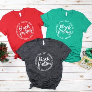 May include: Three t-shirts in red, green, and gray with a white circle design that says "COFFEE &bull; SALES &bull; COUPONS &bull; LUNCH &bull; SHOPPING &bull; MESSY BUN &bull; YOGA PANTS" around the words "black friday".