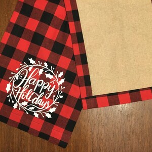 May include: Red and black buffalo plaid table runner with a white embroidered wreath that says "Happy Holidays". The runner has a burlap center panel.