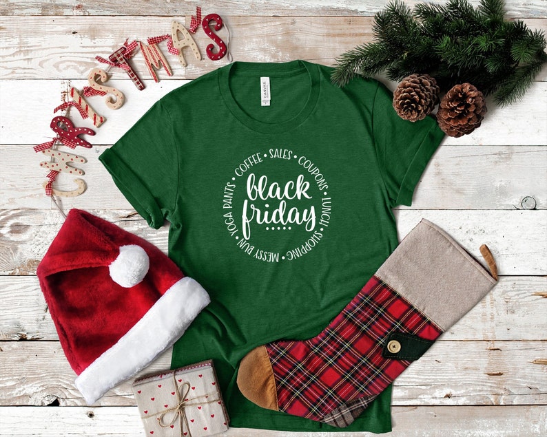 May include: Green t-shirt with a white circle graphic that says "black friday" with a list of activities around the text. The shirt is on a white wood background with a red Santa hat, a small gift box, and a red and black plaid stocking.