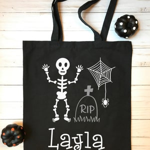 Puede incluir: Bolso tote negro con un esqueleto blanco, telaraña, lápida y el nombre "Layla" impreso. El bolso tiene asas negras y gráficos temáticos de Halloween.