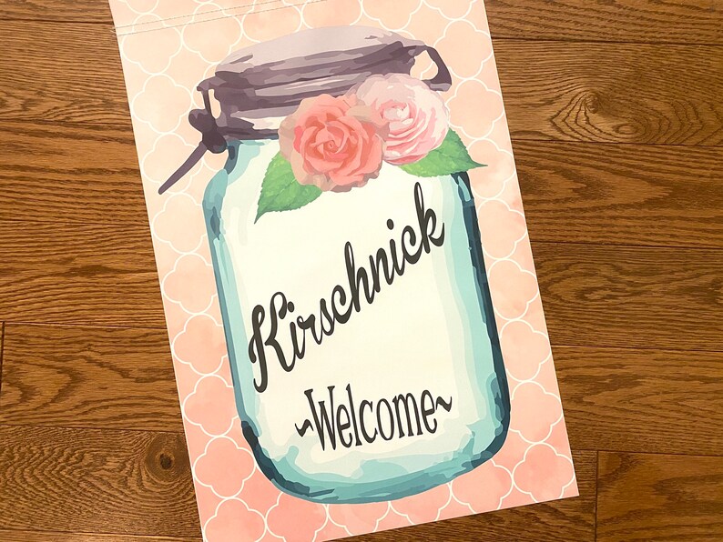 May include: A blue mason jar with a white background and pink flowers on top. The jar has the text "Kirschnick" and "Welcome" written on it. The background is a pink and white pattern.