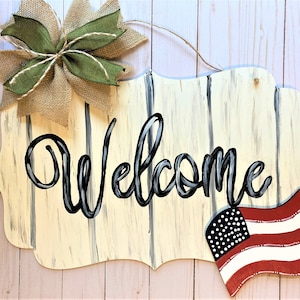 May include: A wooden welcome sign with a distressed white paint finish and black lettering. The sign has a rustic, farmhouse style and features a small American flag painted on the bottom right corner.