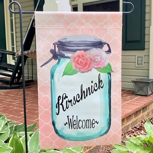May include: A pink and white welcome garden flag with a blue mason jar with flowers and the text "Kirschnick -Welcome-"