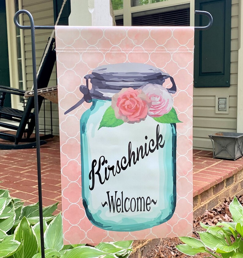 May include: A pink and white welcome garden flag with a blue mason jar with flowers and the text "Kirschnick -Welcome-"