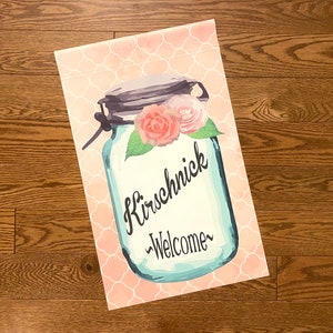 May include: A pink and white welcome garden flag with a mason jar design. The jar has a blue background with the text "Kirschnick -Welcome-" in black. The jar is decorated with pink and white flowers.