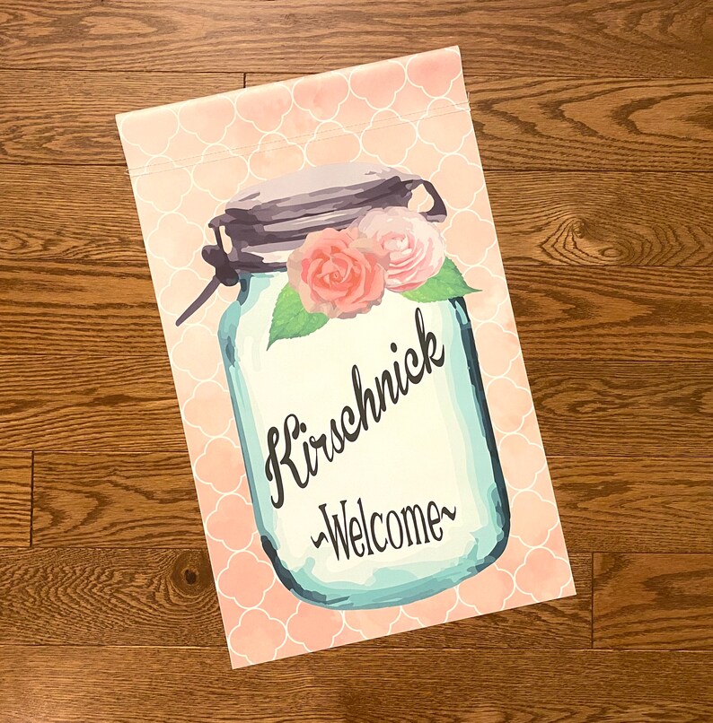 May include: A pink and white welcome garden flag with a mason jar design. The jar has a blue background with the text "Kirschnick -Welcome-" in black. The jar is decorated with pink and white flowers.
