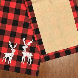 Puede incluir: Un camino de mesa de cuadros rojos y negros con un diseño bordado blanco de dos renos. Un reno tiene el texto "Happy Holidays".
