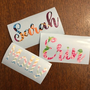 Op de afbeelding: Drie lichtblauwe stickers met namen in kleurrijke, patroonletters. De namen zijn "Sarah" in oranje, blauw en paars, "Erin" in roze bloemen en "Molly" in pastelkleurige zigzagpatronen. Op een houten oppervlak.