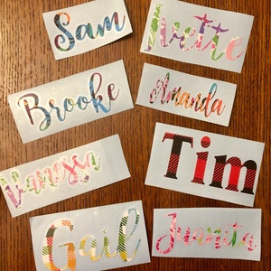 Op de afbeelding: Een verzameling naamstickers in verschillende kleuren en patronen. Namen zijn onder meer "Sam", "Ivettte", "Brooke", "Amanda", "Vanessa", "Tim", "Gail" en "Juanita". De stickers hebben een lichtblauwe achtergrond.