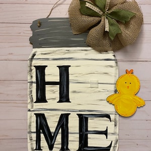 May include: A white wooden mason jar with the word "HOME" painted in black. A burlap bow with a green ribbon is attached to the top of the jar. A yellow chick cutout is attached to the right side of the jar.