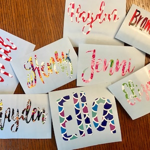 Op de afbeelding: Een verzameling gepersonaliseerde naamstickers op lichtblauwe achtergronden. De namen zijn in verschillende lettertypen en kleuren, waaronder bloemen-, polkadot- en patroonontwerpen. De namen zijn onder meer Hayden, Jenni en Ella.