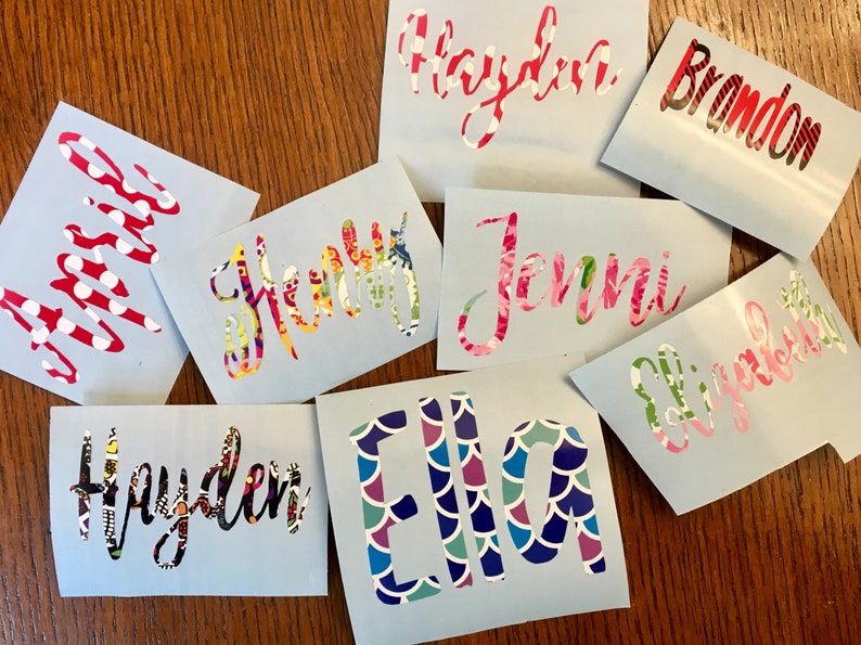 Op de afbeelding: Een verzameling gepersonaliseerde naamstickers op lichtblauwe achtergronden. De namen zijn in verschillende lettertypen en kleuren, waaronder bloemen-, polkadot- en patroonontwerpen. De namen zijn onder meer Hayden, Jenni en Ella.