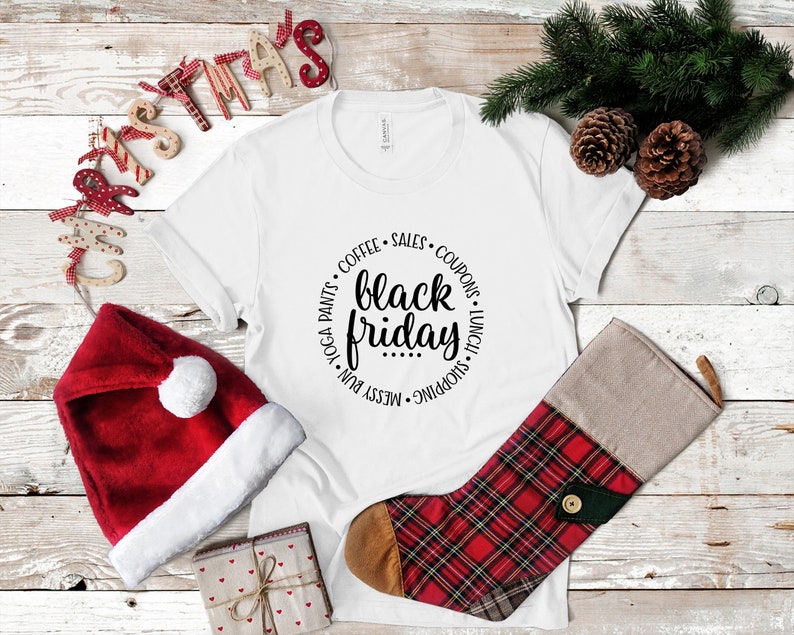 May include: White t-shirt with a black circle graphic that says "messy bun, yoga pants, coffee, sales, coupons, shopping, lunch, black friday". The t-shirt is on a white wood background with a red and white Santa hat and a red and black plaid stocking.