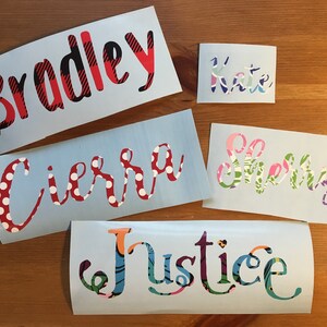 Op de afbeelding: Een verzameling gepersonaliseerde naamstickers in verschillende lettertypen en patronen. De namen zijn onder meer "Bradley" in rood en zwart geruit, "Kate" in pastelkleuren, "Cierra" met rode stippen, "Sherry" met bloempatronen en "Justice" in een kleurrijk ontwerp.