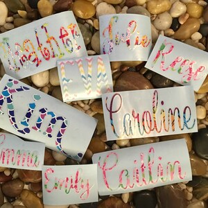Op de afbeelding: Een verzameling gepersonaliseerde naamstickers in verschillende lettertypen en kleurrijke patronen. Namen zijn onder meer "Heather", "Julie", "Reese", "Kim", "Eva", "Caroline", "Caitlin", "Emma" en "Emily". De stickers staan op een lichtblauwe achtergrond.