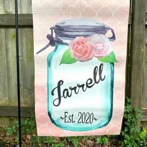 May include: A pink and white patterned garden flag with a blue mason jar with a gray lid and a floral design. The jar has the text "Farrell ~Est. 2020~" on it.