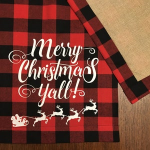 Puede incluir: Un camino de mesa de cuadros rojos y negros con texto blanco que dice "Merry Christmas Y'all!" y una silueta blanca de Papá Noel en un trineo tirado por renos.