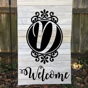 May include: A white wooden welcome sign with a black letter "V" in a decorative circle and the word "Welcome" written in black script.