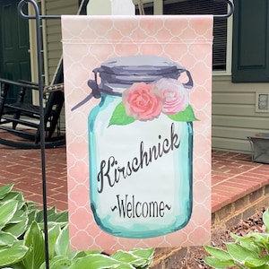 May include: A pink and white welcome garden flag with a blue mason jar with flowers and the text "Kirschnick ~Welcome~".