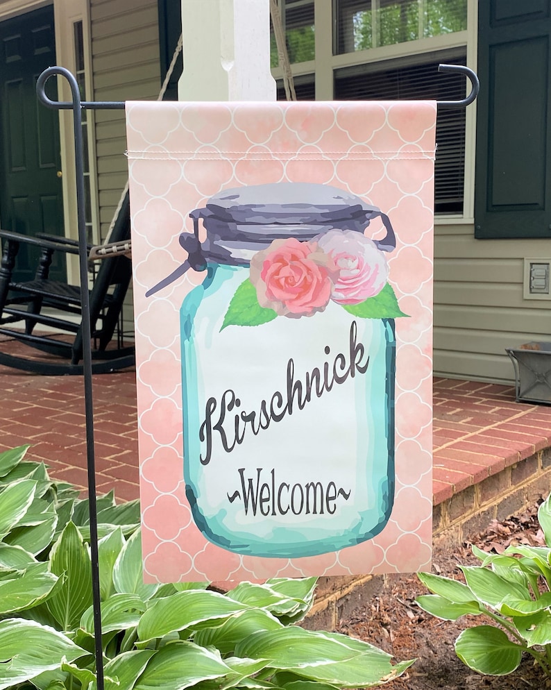 May include: A pink and white welcome garden flag with a blue mason jar with flowers and the text "Kirschnick ~Welcome~".