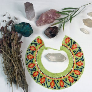 Puede incluir: Un collar de cuentas verdes con detalles en naranja, rojo y azul. El collar está colocado sobre una superficie blanca con varios cristales y hierbas secas.