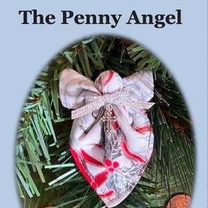 May include: A white fabric angel ornament with red and silver accents, hanging on a green artificial Christmas tree. The angel has a small pocket and a cross charm. The text "The Penny Angel" is at the top of the image. The text below the image reads "Stitch a penny angel from charm squares or quilt scraps. Place a heartfelt message inside the pocket with a penny and spread message of kindness throughout the year!"