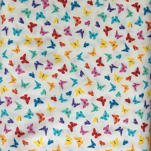 May include: White fabric covered in a repeating pattern of colorful butterflies. The butterflies are in shades of red, yellow, blue, purple, orange, and green. The fabric is likely cotton or a similar material.