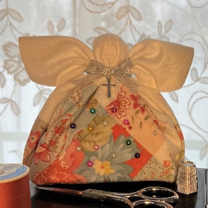 Puede incluir: Un ángel de tela blanca con un acabado brillante y un dije de cruz. El ángel sostiene una bolsa de tela de retazos con patrones florales y cuentas de colores.