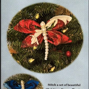 Peut inclure: Ornement de Noël en forme de libellule. La libellule a des ailes rouges et blanches avec des accents dorés, un corps perlé, et est placée sur un fond de branches de sapin. Le texte dit "The Christmas Dragonfly".